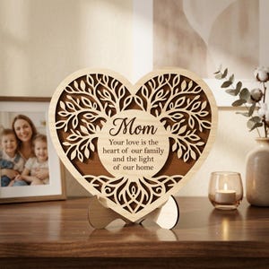 Puede incluir: Un adorno de madera en forma de corazón con un diseño de árbol y la palabra "Mamá". El texto dice: "Tu amor es el corazón de nuestra familia y la luz de nuestro hogar". Se apoya en una base de madera.