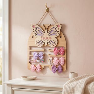 May include: Wooden butterfly-shaped hair bow holder with the name "Emma" engraved. The holder has slots for bows and clips, and is adorned with pink, purple, and light pink bows. It hangs from a gold hook on a pink wall.