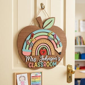 May include: Wooden apple-shaped classroom sign with a rainbow design, pencil, and ruler. The sign includes the text "Mrs. Johnson's Classroom" in colourful letters. Paper clips are attached to the rainbow. The sign hangs on a door.