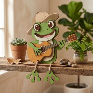 Pode incluir: Estatueta de madeira de um sapo tocando guitarra. O sapo é verde com um chapéu castanho e um rosto sorridente. A guitarra é castanha e bege. As pernas do sapo estão penduradas. À esquerda, há uma pequena planta suculenta.