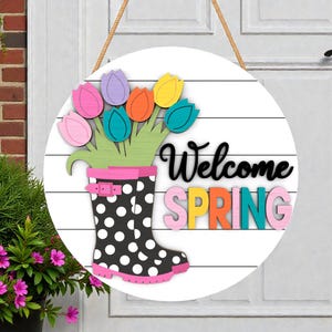 May include: A round wooden sign with a white background and horizontal wood grain. It features a black and white polka dot rain boot with a pink trim, holding colorful tulips. The words "Welcome SPRING" are displayed in black and colorful letters.