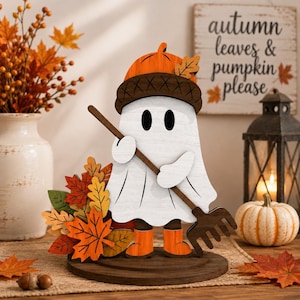 Può includere: Una decorazione da tavolo in legno raffigurante un fantasma bianco con un cappello a forma di ghianda arancione e stivali arancioni, che tiene in mano un rastrello marrone. Il fantasma poggia su una base circolare marrone accanto a foglie autunnali.