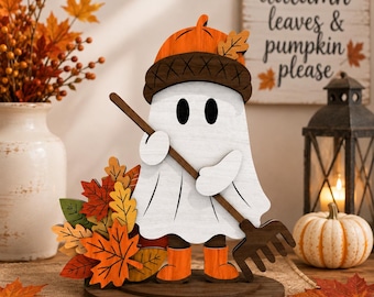 May include: A wooden tabletop decoration featuring a white ghost wearing an orange acorn hat and orange boots, holding a brown rake. The ghost stands on a circular brown base next to a cluster of colorful autumn leaves in shades of red, orange, and yellow.