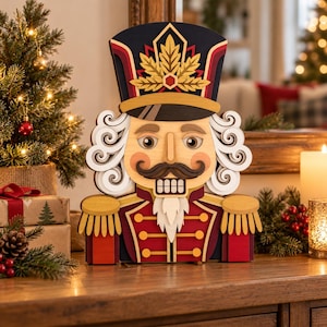 Peut inclure: Une décoration de casse-noisette en bois superposé avec un chapeau bleu foncé orné d'une feuille d'érable dorée, un uniforme rouge aux détails dorés, des cheveux blancs bouclés, une moustache brune et une barbe blanche.