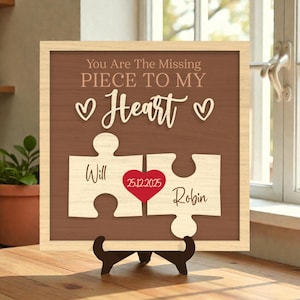 Puede incluir: Letrero de madera con diseño de pieza de rompecabezas, con el texto "You Are The Missing Piece To My Heart". Incluye los nombres "Will" y "Robin" en piezas de rompecabezas, un corazón rojo con la fecha 25.12.2025. El letrero se muestra en un soporte negro.