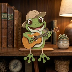 Peut inclure: Figurine en bois repr&eacute;sentant une grenouille verte avec un chapeau et une guitare. La figurine est pos&eacute;e sur une &eacute;tag&egrave;re en bois, avec des livres et une petite plante en pot en arri&egrave;re-plan.