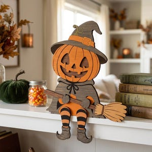 Può includere: Una decorazione in legno intagliato raffigurante una figura con testa di zucca, cappello da strega e mantello, che tiene in mano una scopa. La figura ha gambe a righe arancioni e nere e stivali a punta, progettata per sedersi su un bordo.