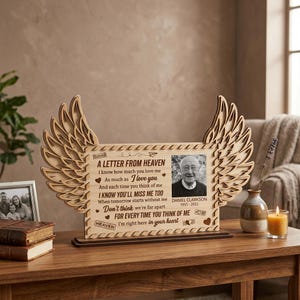 May include: A wooden memorial photo frame with angel wings. The frame features a black and white photo of a man and the text "A Letter From Heaven." The frame is on a wooden table with other decorative items.