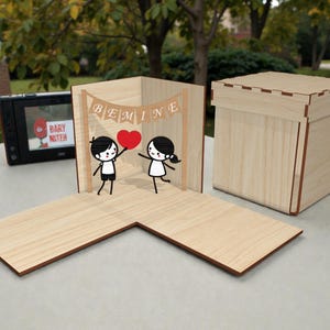May include: A wooden pop-up card featuring stick figures and a heart, with the word "BEMINE" on a banner. A matching wooden box is also visible. The card is open, and the box is closed.