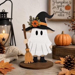 Peut inclure: Une figurine de fantôme en bois portant un chapeau de sorcière noir orné de fleurs orange et de feuilles vertes. Le fantôme est blanc avec des yeux ovales noirs, des bottes noires et tient un balai marron clair sur un socle circulaire en bois.