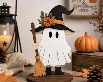 May include: A wooden ghost figurine wearing a black witch hat adorned with orange flowers and green leaves. The ghost is white with black oval eyes, black boots, and holds a light brown broom. It stands on a circular dark brown wooden base.