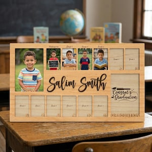 May include: A wooden photo frame with spaces for school photos from Pre-K to 11th grade. The frame includes the name "Salim Smith" and the text "Congrat's Graduation". The frame is made of light-colored wood and sits on a wooden desk.
