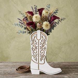 Puede incluir: Un florero de madera blanca con forma de bota vaquera que presenta intrincados patrones cortados con láser. El florero contiene un arreglo floral seco compuesto por calas color burdeos, flores crema, ramas de lavanda y hojas de eucalipto.