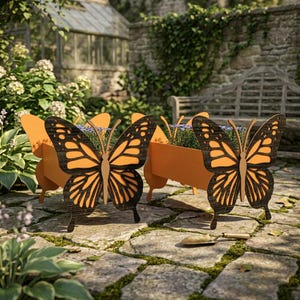 May include: Orange butterfly-shaped planters with black wing details, set on a stone patio. The planters have rectangular boxes for flowers and are in a garden setting with a greenhouse and bench in the background.