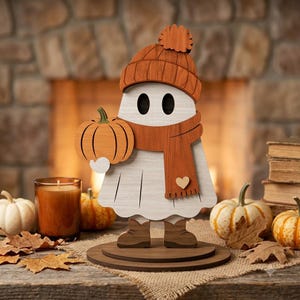 Könnte beinhalten: Eine weiße Geisterfigur aus Holz mit einer orangefarbenen Strickmütze mit Bommel und einem passenden orangefarbenen Schal mit einem kleinen Herzdetail. Der Geist hält einen kleinen orangefarbenen Kürbis und steht auf einem runden Holzsockel, bekleidet mit braunen Stiefeln.