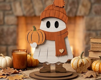 May include: A white wooden ghost figurine wearing an orange knit-style hat with a pom-pom and a matching orange scarf with a small heart detail. The ghost holds a small orange pumpkin and stands on a circular wooden base, wearing brown boots.