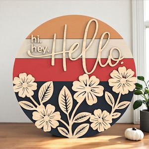 Könnte beinhalten: Runder Holzschild mit dem Wort "Hello" in Schreibschrift, überlagert von einem Design mit orangefarbenen, weißen, roten und marineblauen Streifen. Das Schild zeigt auch lasergeschnittene Blumen und die Worte "hi. hey."