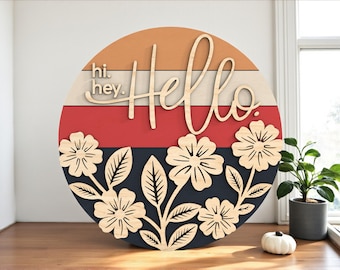 May include: A round wooden sign with the word "Hello" in cursive, layered over a design with orange, white, red, and navy blue stripes. The sign also features laser-cut flowers and the words "hi. hey."