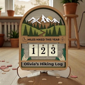 May include: Wooden hiking log with a mountain scene, a compass, and the text "Miles Hiked This Year." The log displays the numbers "123" and the name "Olivia's Hiking Log."