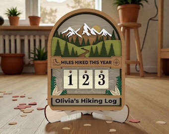 May include: Wooden hiking log with a mountain scene, a compass, and the text "Miles Hiked This Year." The log displays the numbers "123" and the name "Olivia's Hiking Log."
