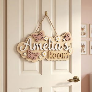 Può includere: Un cartello in legno per la porta della stanza con la scritta "Amelia's Room" in corsivo bianco. Il cartello è decorato con farfalle rosa e margherite bianche. È appeso a una corda di iuta su una porta bianca.