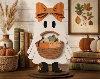 May include: A white wooden ghost cutout decoration featuring a large orange bow on its head and black boots. The ghost holds a brown woven basket filled with three small wooden pumpkins in shades of orange, green, and cream, standing on a base.