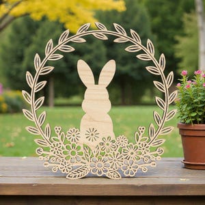 Peut inclure: Une couronne de Pâques en bois avec un motif floral et de feuilles. Le lapin est au centre de la couronne. Une plante en pot avec des fleurs roses est à droite de la couronne. La couronne est sur une surface en bois.