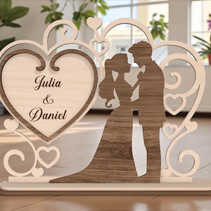 Könnte beinhalten: Holz-Hochzeitsdekoration mit einer Silhouette von Braut und Bräutigam, mit den Namen "Julia & Daniel" auf einem Herzen graviert. Hergestellt aus hellem und dunklem Holz, mit dekorativen Herzakzenten.