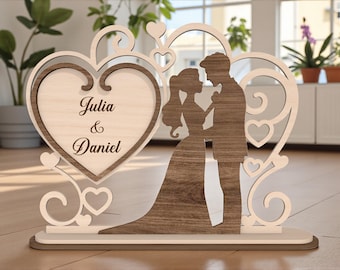 Può includere: Decorazione nuziale in legno con silhouette di sposi e i nomi "Julia & Daniel" incisi su un cuore. Realizzato in legno chiaro e scuro, con dettagli decorativi a forma di cuore.