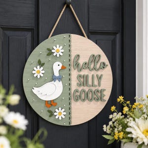 May include: A round wooden door sign with a painted green and natural wood finish. The sign features a white goose with an orange beak and feet, a blue bow tie, and daisy accents. The words "hello SILLY GOOSE" are in green.