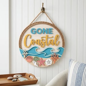 Peut inclure: Panneau rond en bois avec l'inscription "GONE Coastal" en bleu et jaune. Le panneau représente une scène de plage avec des vagues, des coquillages et du corail. Il est suspendu à un crochet sur un mur blanc.