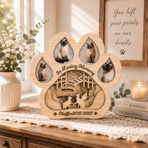May include: A light-colored wooden paw-shaped memorial frame featuring three oval photo slots, the text In Loving Memory, and a layered cutout design of two cats with halos and wings. The base is engraved with Muffin 2012-2021 and paw prints.