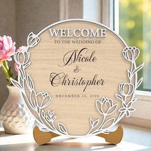 Pode incluir: Uma placa de boas-vindas de casamento em madeira com os nomes Nicole & Christopher e a data de 18 de dezembro de 2026. A placa é redonda com um design floral e um suporte de madeira. Um vaso com flores cor-de-rosa está à esquerda.