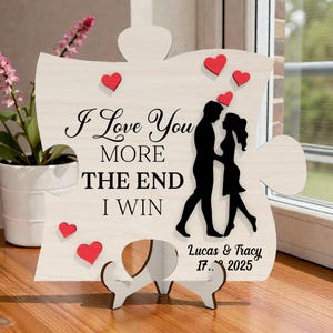 Puede incluir: Una pieza de rompecabezas de madera con las palabras "I Love You More The End I Win" en escritura negra. Presenta una silueta negra de una pareja y acentos de corazón rojo. También se incluyen los nombres "Lucas & Tracy" y la fecha "17.. 2025".