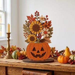 Op de afbeelding: Een decoratief houten jack-o'-lantern middelpunt met een uitgesneden oranje pompoenbasis, bekroond met een gelaagd arrangement van zonnebloemen, herfstbladeren en rode bessen, alles gemonteerd op een ronde donkerbruine houten voet.