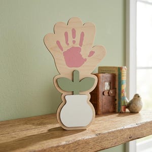 May include: Wooden flower-shaped decoration with a handprint in pink paint. The flower has a light green stem and leaves, and a white vase. The decoration is on a wooden shelf.