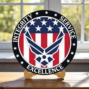 Puede incluir: Un letrero redondo de madera con el emblema de la Fuerza Aérea de los Estados Unidos. El letrero tiene un fondo a rayas rojas y blancas con un emblema azul y estrellas blancas. Las palabras "INTEGRITY SERVICE EXCELLENCE" están en el borde.