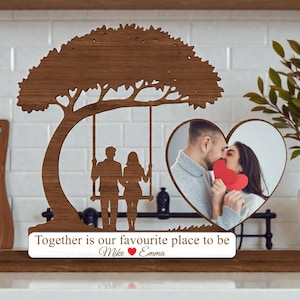 May include: A wooden photo frame with a tree design, a swing with silhouettes of a couple, and a heart-shaped photo. The frame includes the text "Together is our favourite place to be" and names. The photo shows a couple kissing.