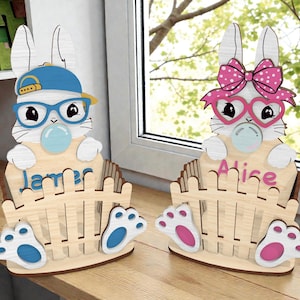 May include: Two wooden Easter bunny decorations. One bunny wears a blue cap and glasses, blowing a bubble, with the name "James". The other bunny has a pink bow and heart-shaped glasses, with the name "Alice". Both are in front of a wooden fence.