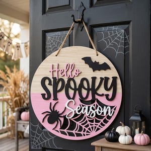 Puede incluir: Un letrero de puerta redondo de madera con el texto Hello Spooky Season en letras negras, rosas y blancas. El letrero incluye una silueta de murciélago negro, una araña negra y un diseño de telaraña, suspendido por una cuerda.