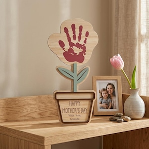 Puede incluir: Decoración floral de madera con una huella de mano en la flor. La flor está en una maceta con el texto "HAPPY MOTHER'S DAY With love, Amanda". Un marco de fotos y un jarrón con un tulipán rosa también están sobre la superficie de madera.