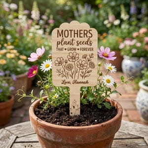 Pode incluir: Uma estaca de jardim de madeira com o texto "Mothers plant seeds that grow forever" e um design floral. A estaca está num vaso de terracota com flores. O texto "Love, Hannah" está na parte inferior.