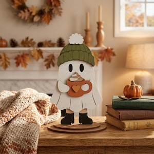 May include: A white wooden ghost figurine wearing a green knit-style hat with a white pom-pom and black boots. The ghost holds an orange mug with a heart detail and a swirl of whipped cream on top, standing on a circular wooden base.