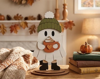 Puede incluir: Una figura de fantasma de madera blanca con un gorro de punto verde con un pompón blanco y botas negras. El fantasma sostiene una taza naranja con un detalle de corazón y una espiral de nata montada, sobre una base circular de madera.