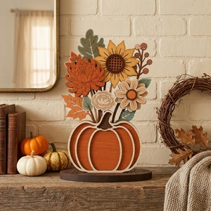Può includere: Una decorazione da tavolo in legno a strati con una base a forma di zucca arancione intagliata che sostiene un bouquet di fiori e foglie ritagliate nei toni del giallo, arancione, crema e verde salvia, montata su una base circolare in legno scuro.