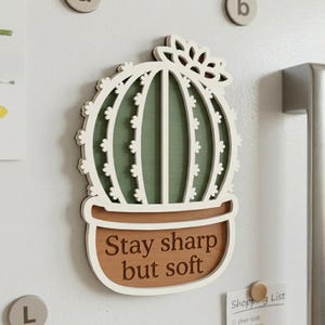 May include: A wooden cactus-shaped magnet with a white outline and green interior. The magnet has the text "Stay sharp but soft" in a brown pot. The magnet is attached to a white surface.