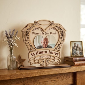 Könnte beinhalten: Ein hölzerner Gedenk-Fotorahmen mit herzförmigem Ausschnitt, Engelsflügeln und dem Text "Always on our minds, Forever in our hearts". Der Rahmen enthält ein Foto eines Mannes und den Namen "William Jones 1954-2020."