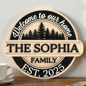 Puede incluir: Un letrero redondo de madera con el texto "Welcome to our home" y "The Sophia Family EST. 2025". El letrero presenta un esquema de color negro y madera natural con un diseño de silueta de bosque. Un artículo decorativo para el hogar.