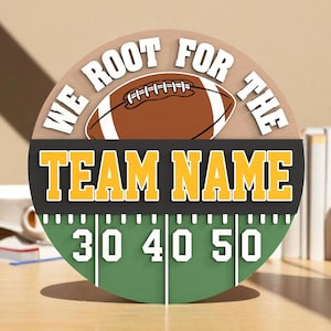 May include: A round wooden sign with a football theme. The sign features a football graphic and the text "WE ROOT FOR THE TEAM NAME" in white and yellow. The bottom half of the sign has a green field with yard lines and the numbers 30, 40, and 50.