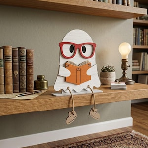 Op de afbeelding: Een wit houten spookfiguur met een rode bril die een oranje boek vasthoudt. Het spook heeft twee bungelende benen van touw met houten voeten eraan vast. Het figuur is ontworpen om op een plat oppervlak te zitten met de benen over de rand.